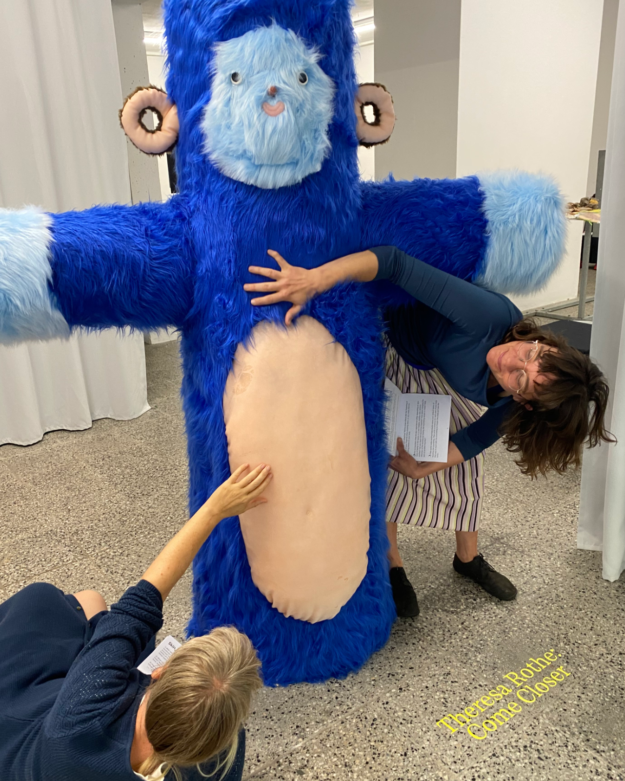 The photo shows two women touching a human sized fluffy figure, which is one of the artworks by Theresa Rothe displayed in the exhibition at Leipzig Kunstverein. The figure has blue fur, and a tiny face that feels sticky to the touch. The woman on the right is beding her body so that she fits underneath the figure’s voluminous arm. Her own arm comes up, and touches the figures fur above its belly. The other woman is lying on the ground, and reaching upwards to feel the leathery belly of the creature. Both women are holding a Tactile Descriptions Workbook booklet in the hand that is not touching the figure. The mood is curious and playful.