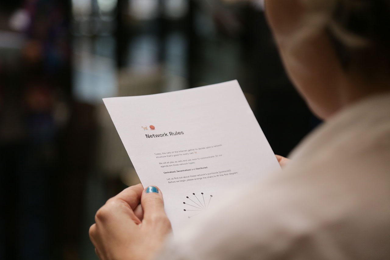 The photo is taken over the shoulder of a workshop participant who is holding the instructions for the physical game in their hands. Only the headline, Network Rules, can be read, with two emojis of a cat and a call of yarn printed above.