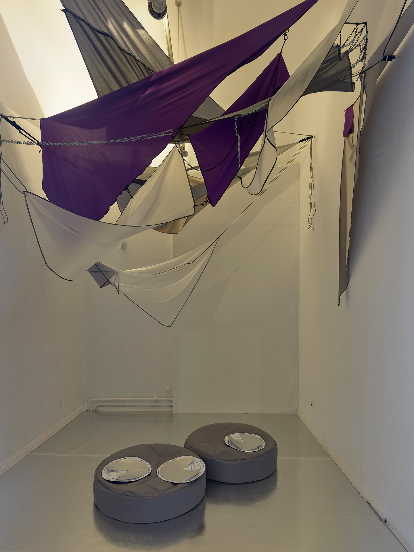 Room with geometric fabric art installation in shades of purple, gray, and beige suspended from the ceiling. The fabric pieces each stand for a star costellation and are held together and hug with various hooks, chains and zippers. Two round gray cushions with shiny silver tops are on the glossy floor below, with star constellations charts atop.