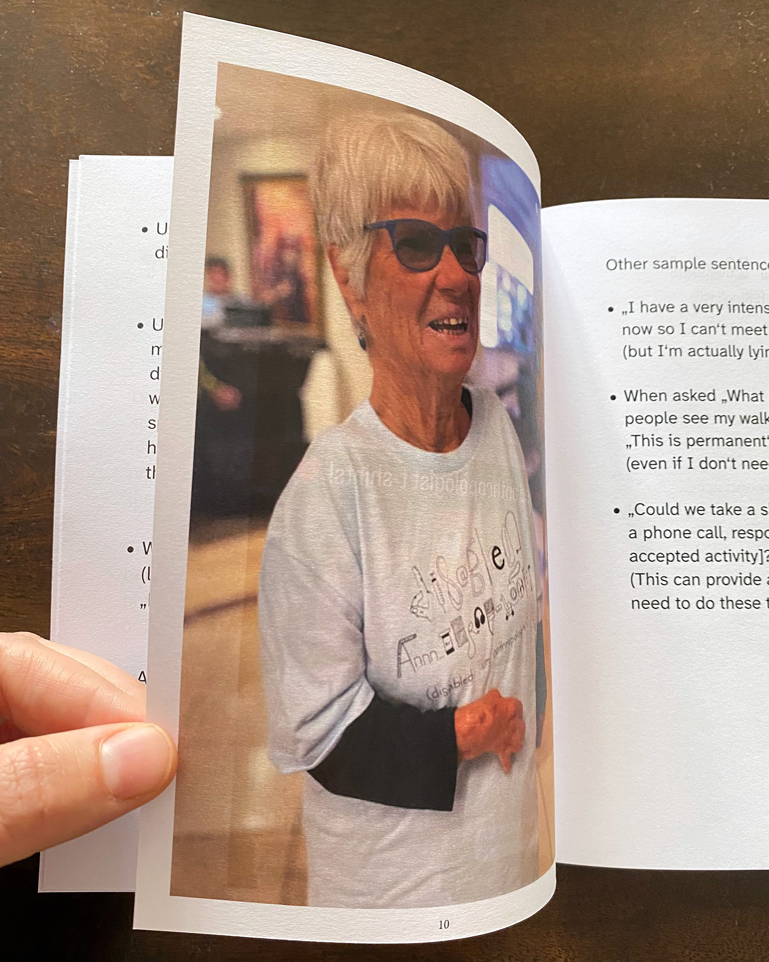 Photo of a hand flipping through the opened booklet on a dark wooden table. The opened page shows a photograph of of white, queer, disabled anthropologist Sumi Colligan who is in her 70s. She is smiling and has short grey hair, blue glasses, and is wearing a long black shirt layered with the light blue disabled anthropologist t-shirt.