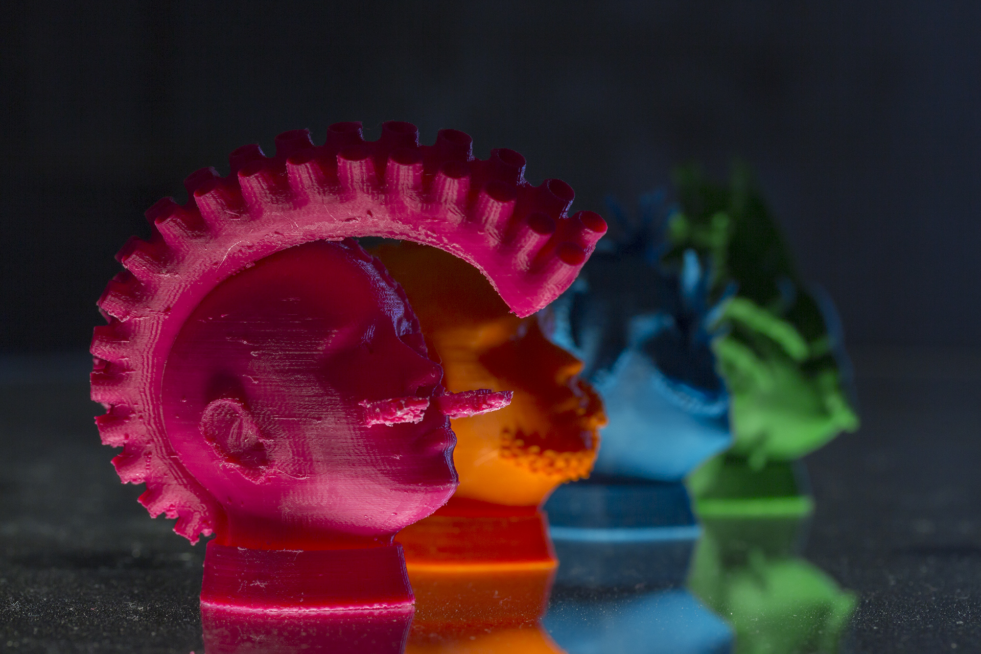 Four 3D-printed heads in pink, organge, blue and green are arranged diagonally on a mirroring surface with the pink head in the front. This head has USB cables sticking out of their cheecks, and a large tentacle instead of hair.