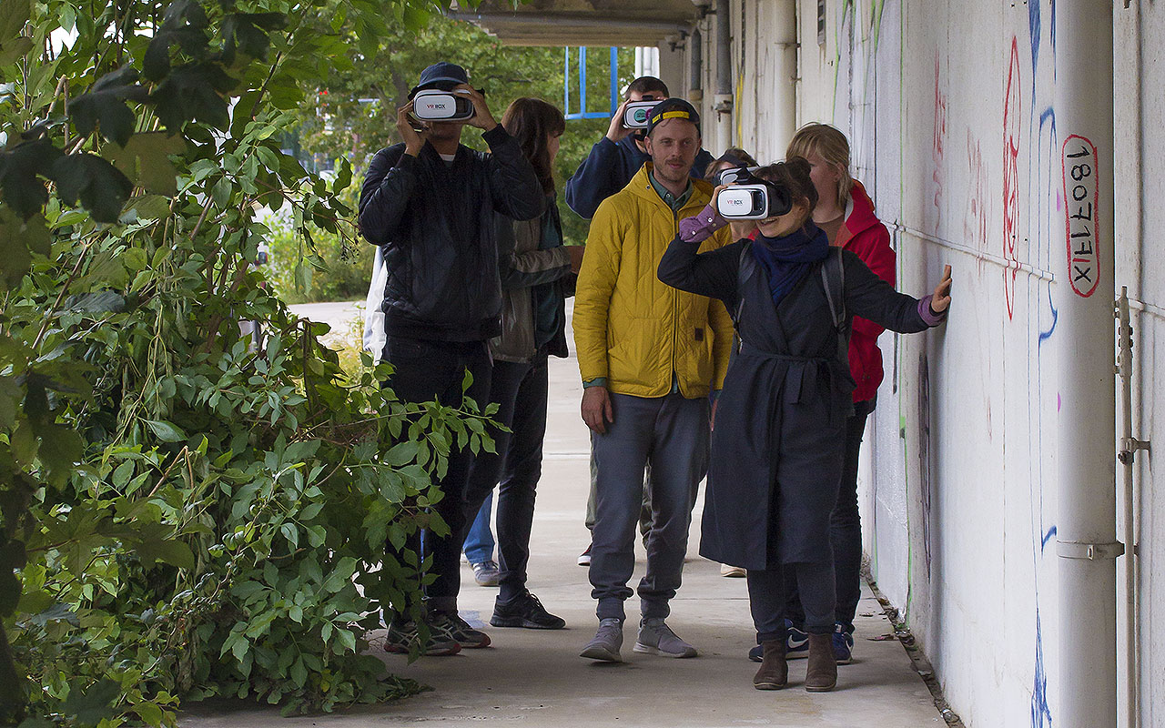 On an Augmented Reality Walk, three out of six participants are wearing white AR headsets. The person in the front is touching the headset with their hand, and using the other hand to touch the concrete wall next to them.