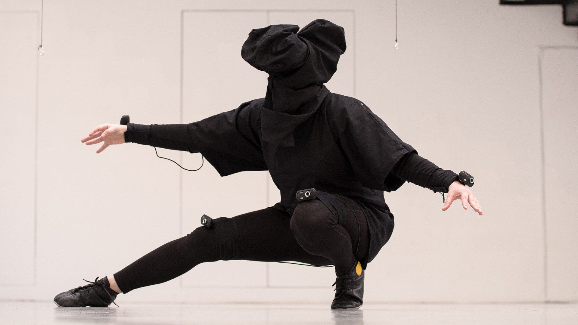 The photo is filled by a person who is wearing a dark, self-made Virtual Reality Suit that consists of a hood for the head in which a VR headset is hidden, a large shirt that has webcams attached to it, and leggins. They are crouched down, with one leg extended and one leg holding their weight. Their arms are reaching towards the sides of the image. Webcams are attached to their wrists, and to both knees.