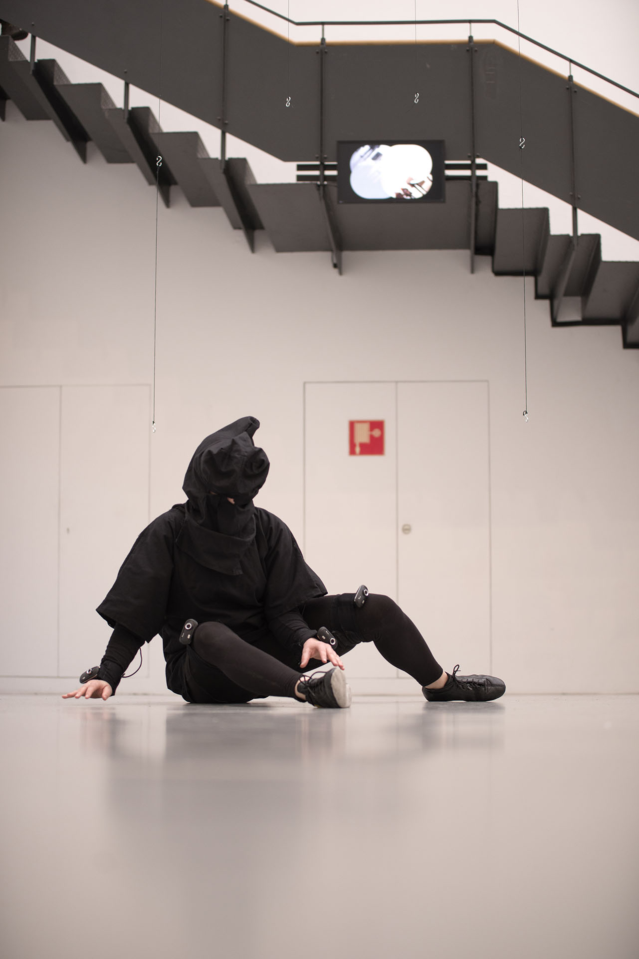 A performer wearing a Virtual Reality suit is sitting on the ground and exploring visual input directed towards a VR headset in a dark hood via webcams that are attached to their wrists and knees. Their legs are turned slightly outwards to the left and their right hand in positioned in between the legs. Above them, a video screen attached to a large staircase shows the four video streams that the performer can see through their headset. These images are blurred in the photo.