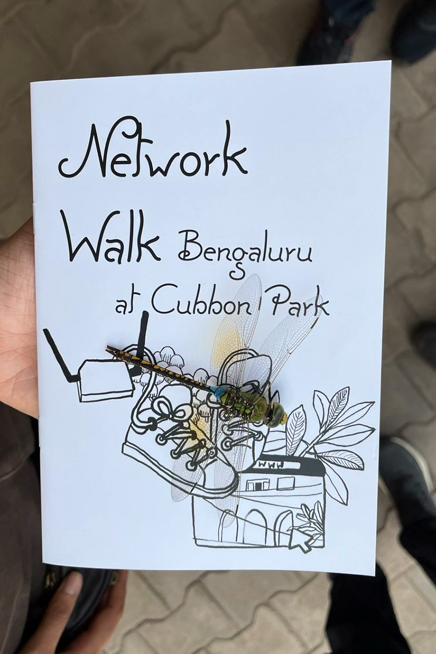 A hand holds the Nework Walk Booklet into the camera. A large dragonfly has landed on the booklet and sits on an illustration of shoes, routers, fungi and leafs.
