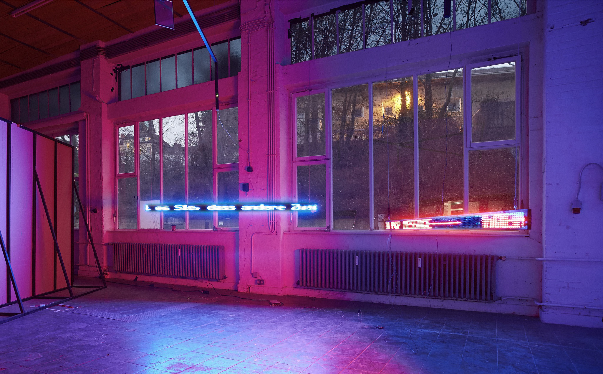 This installation consists of two large LED-boards hanging from the ceiling with one showing an encrypted message in pink and one an decrypted message in blue. The messages themselves are not legible on the photo.