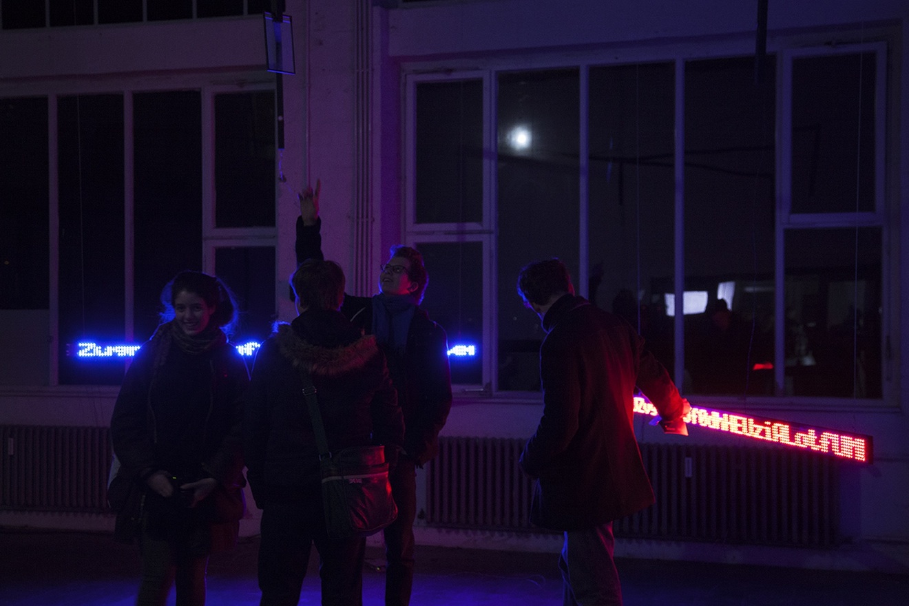 A group of students engage with the installation. Standing between two large LED boards, they point upwards in the direction of a laser.
