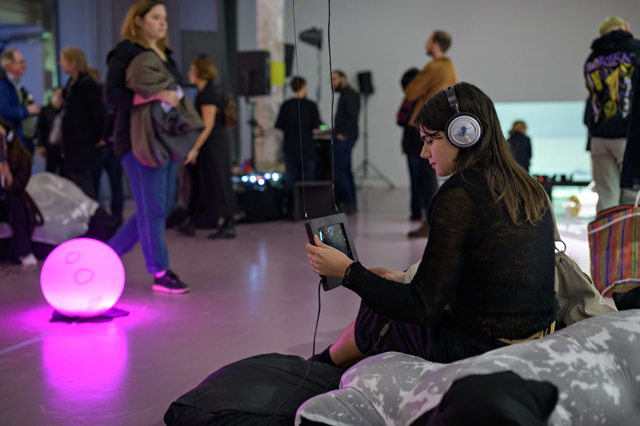 A person with long brown hair sits on a large pillow made from two fabrics, one of them black, one white-silvery transparent with tactile elements underneath its surface. The person is wearing headphones and using one of the tablets that show the website artwork. All around, there are people moving. A pink lamp offers soft pink light.