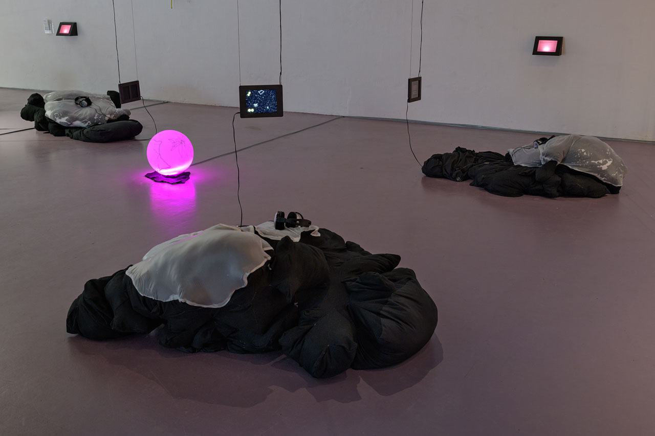 Three large pillows invite listeners to rest and to engage with tablets with a website artwork open on them. In the open space between the pillows, a round lamp sits on the floor with some line drawings on it. The light is pink.
