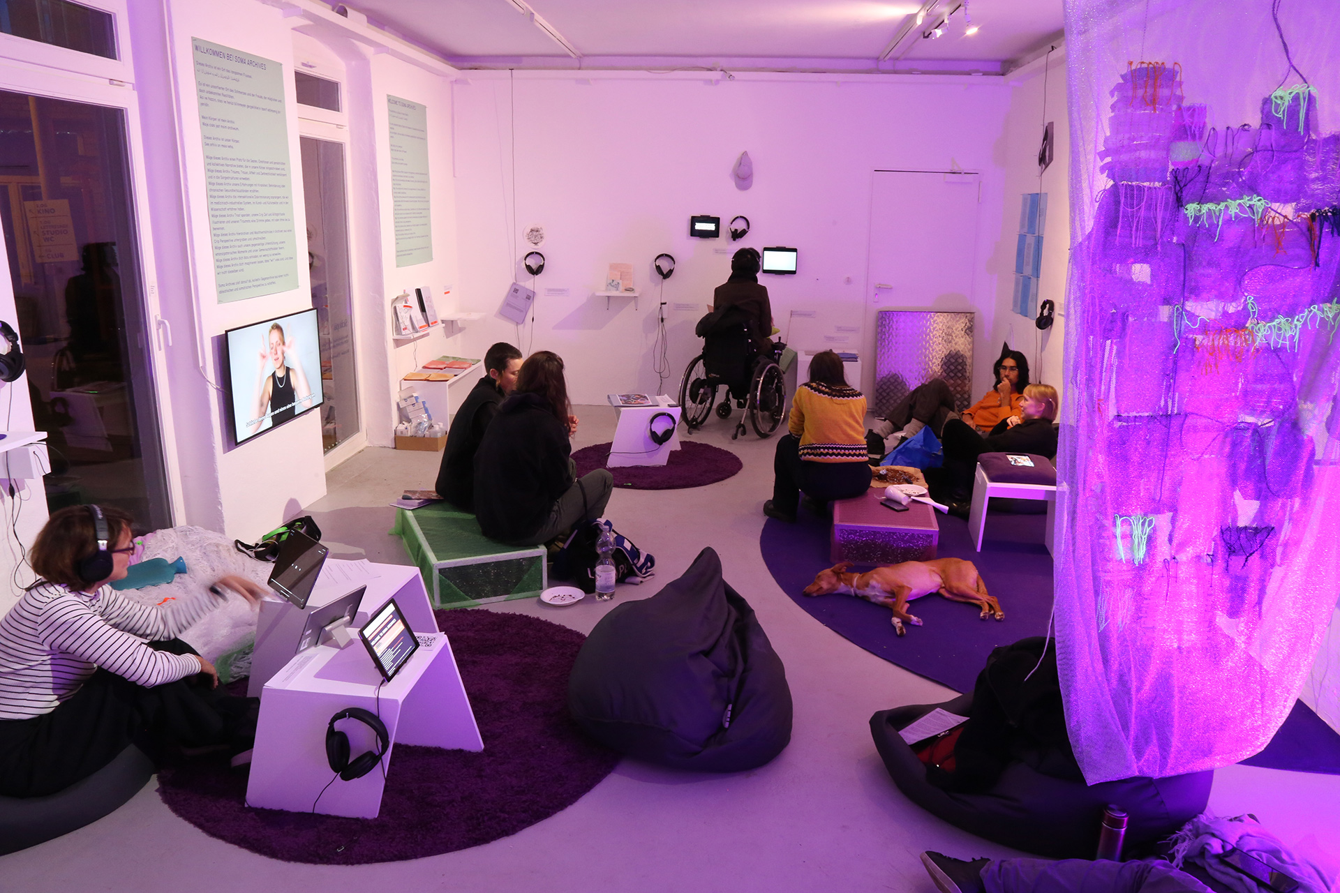 In a cozy exhibition space, multiple people engage with the art works and talk to each other. On the left, there are tablets showing web artworks, and from the right side a violet semitransparent curtain falls softly into the frame. Six people and one dog are sitting and lying on cushions, one person in a wheelchair is watching a film, and the leftovers of a cake are on a table.