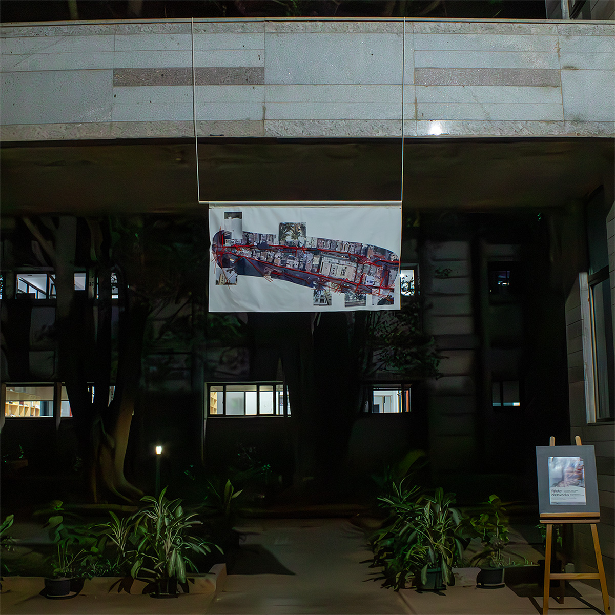 A photo taken during the opening night shows the work Yarn and Wires. The banner is about 1.5m times 0.8 m in dimensions, and has the screenshot of two streets as captured on Google Maps Satellite mode printed across. With red yarn, all cables in the streets are stitched atop. The work is hanging from a concrete bridge in the Southern Laboratories building at the National Center for Biological Sciences Bangalore. Below, plants in pots are positioned around a pathway that one can follow to move underneath and beyond the banner. The darkness in the background is interrupted by the lights of windows.