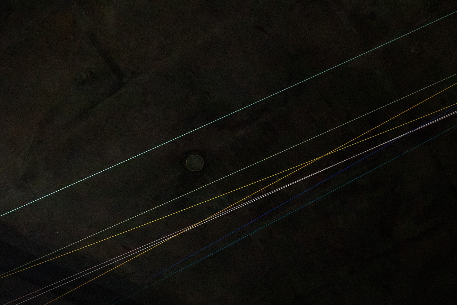 Dark background with six parallel strings running diagonally. Lines are in green, blue, yellow, white, and another blue, intersecting at times. These lines are installed at the ceiling of the exhibition space and represent the length of a hotspot.