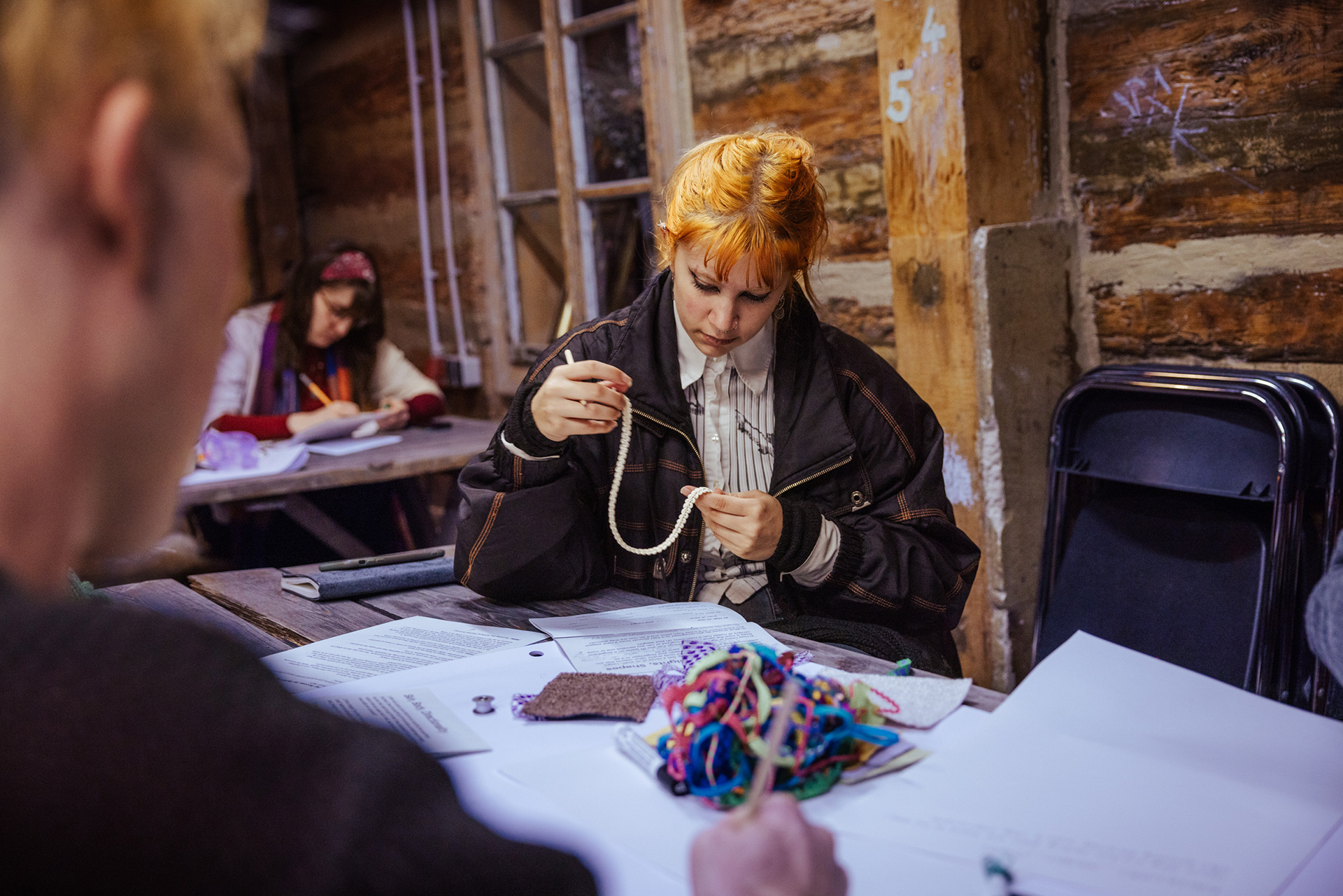 A workshop participant with orange hair tied up in a bun is sitting at a large table full of fabric samples and large pieces of paper. She is moving a piece of fabric through her hands and seems very focused. Two other participants, one sitting opposite and one further back at a different table are writing onto the large pieces of paper.