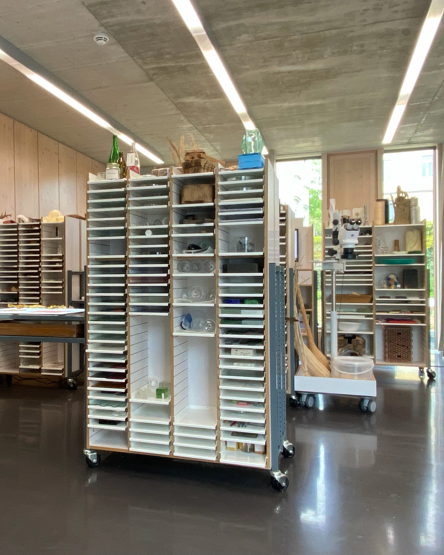 This photo of the material collection shows a large room. The walls are made from wooden panels, the ceiling is made from concrete. The room is filled with large shelves that hold a variety of different materials such as glass, wood, felt, plastics and more.