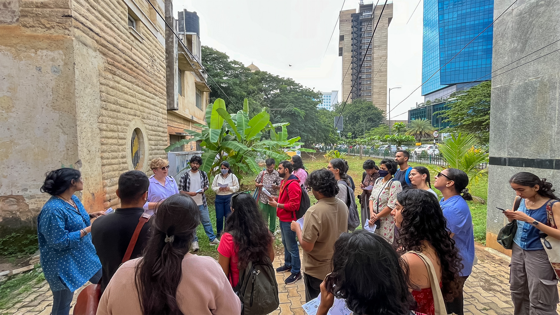 A group of people stand in an outdoor urban area, surrounded by buildings and greenery, with the Central Telegraph Office to their left. They are attentively facing Iz who is talking about the history of the CTO. The setting includes a partially constructed high-rise and trees.