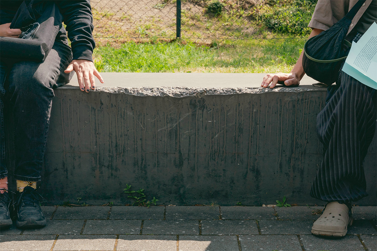 Two people sit on a concrete bench, and each one has one hand reaching into the space between them, where the concrete is cracked open. They are calmly exploring the crack with their fingers.