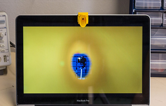 A macbook with the webcamera obscura in the shape of a toilet partially covering its webcam is positioned on a table. On the screen, one can see yellow with an opening in the middle that makes a camera on a tripod visible, since the lid of the toilet is open and lets some webcam footage through.